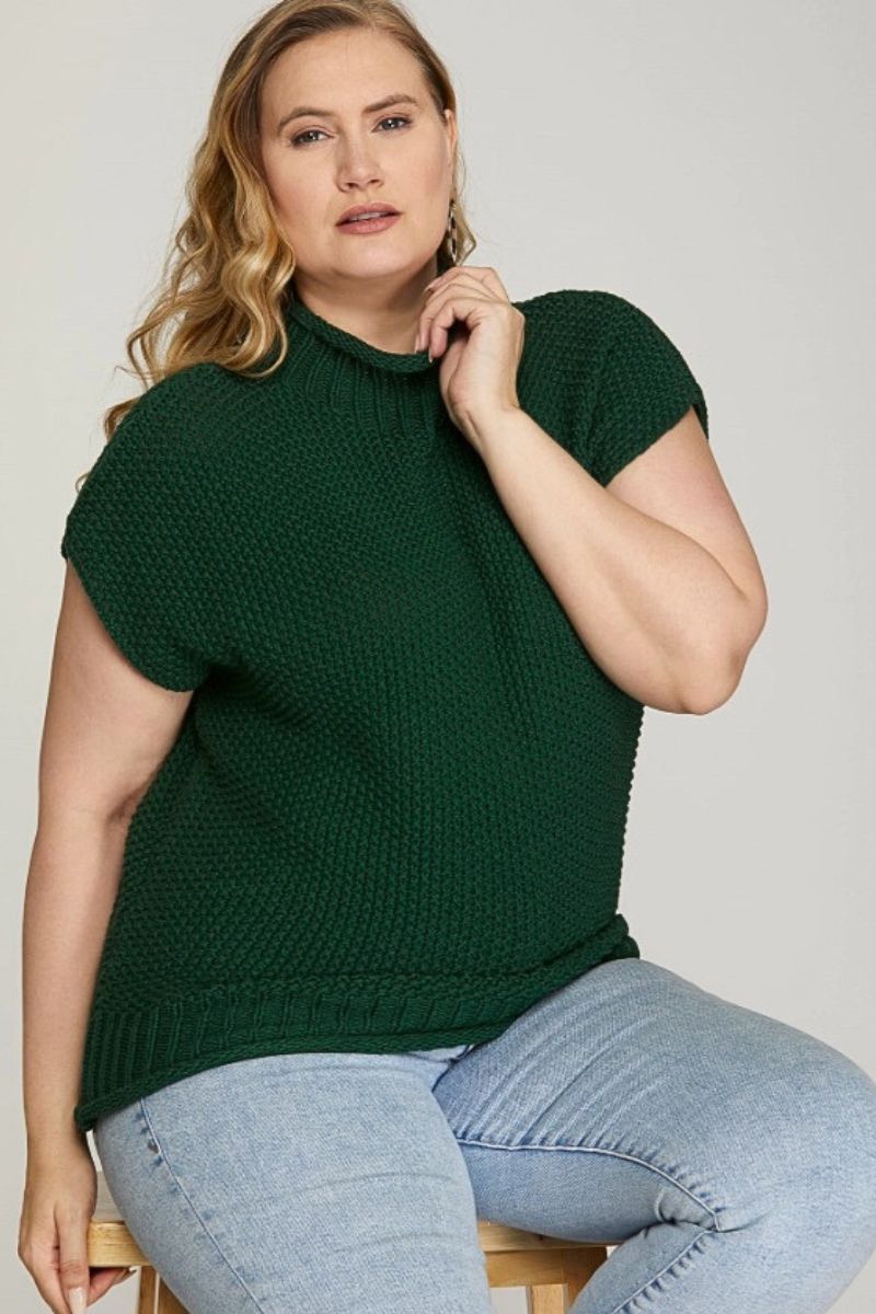 Woman wearing a green knitted top and light blue jeans sitting on a white background